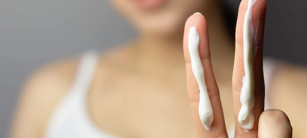 Close-up of a person holding up two fingers, each with a strip of sunscreen. The person is blurred in the background, wearing a white tank top