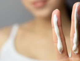 Close-up of a person holding up two fingers, each with a strip of sunscreen. The person is blurred in the background, wearing a white tank top