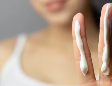 Close-up of a person holding up two fingers, each with a strip of sunscreen. The person is blurred in the background, wearing a white tank top