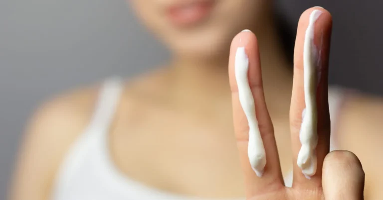 Close-up of a person holding up two fingers, each with a strip of sunscreen. The person is blurred in the background, wearing a white tank top