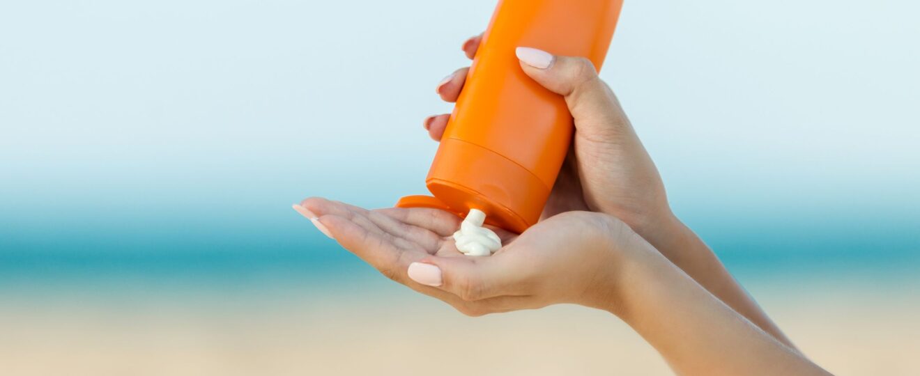 Close-up of hands squeezing mineral sunscreen from an orange bottle onto the palm, with a blurred beach and ocean background.