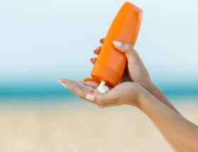 Close-up of hands squeezing mineral sunscreen from an orange bottle onto the palm, with a blurred beach and ocean background.