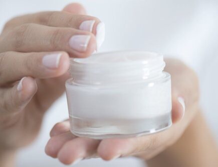 A person's hands holding and applying a small amount of white moisturiser from an open jar.