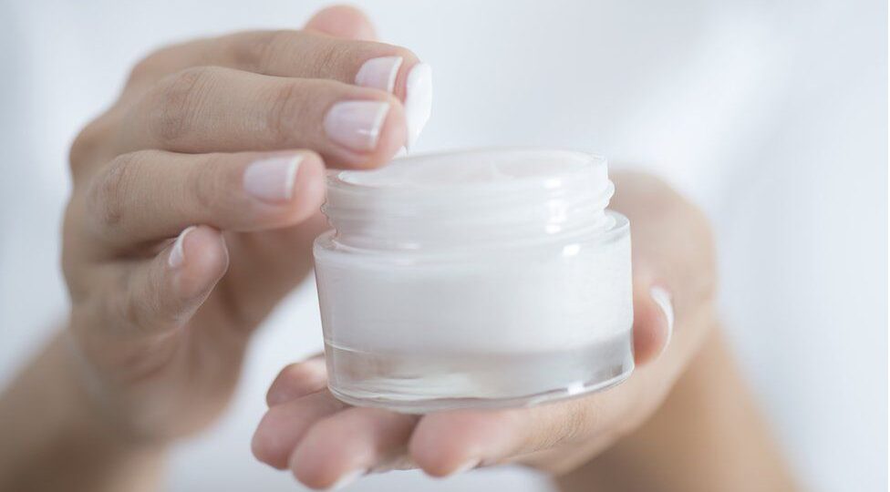 A person's hands holding and applying a small amount of white moisturiser from an open jar.