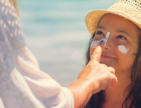 An adult applying SPF 50 sunscreen to a child's face at the beach. The child is smiling and wearing a straw hat, with visible sunscreen on their cheeks and nose.
