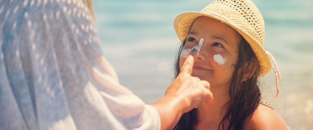 An adult applying SPF 50 sunscreen to a child's face at the beach. The child is smiling and wearing a straw hat, with visible sunscreen on their cheeks and nose.